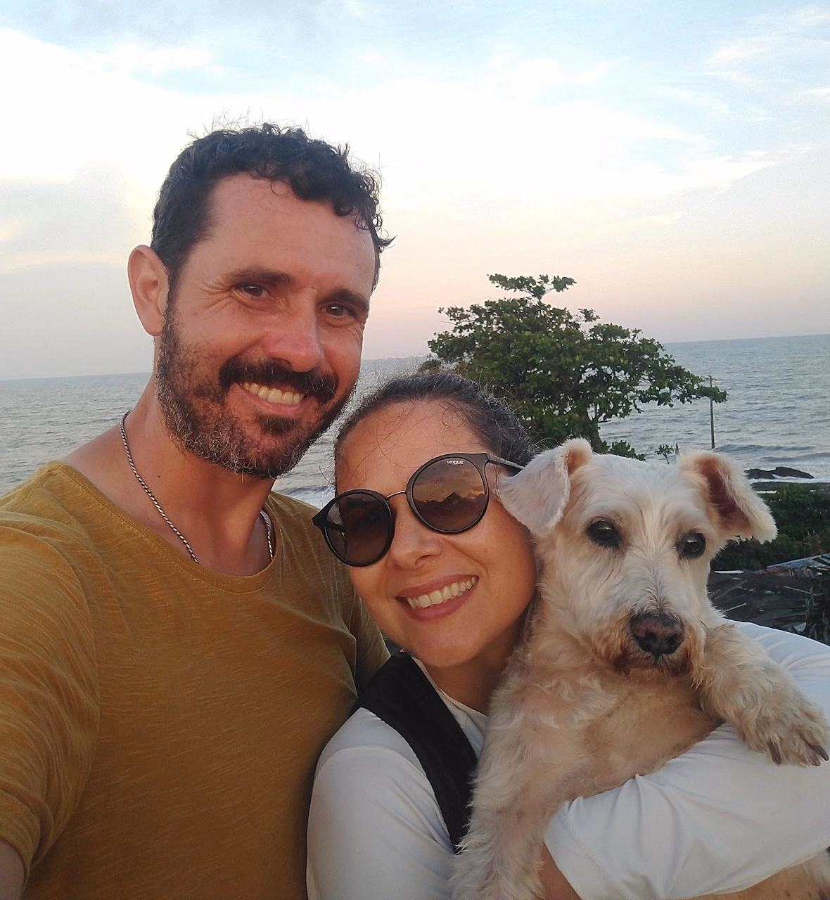 Foto tirada no mirante de Matinhos mostrando eu, a Ju e o Vígi. O Vígi está nos braços da Ju e eu ao lado dos dois. Ao fundo vemos o mar e uma árvore.